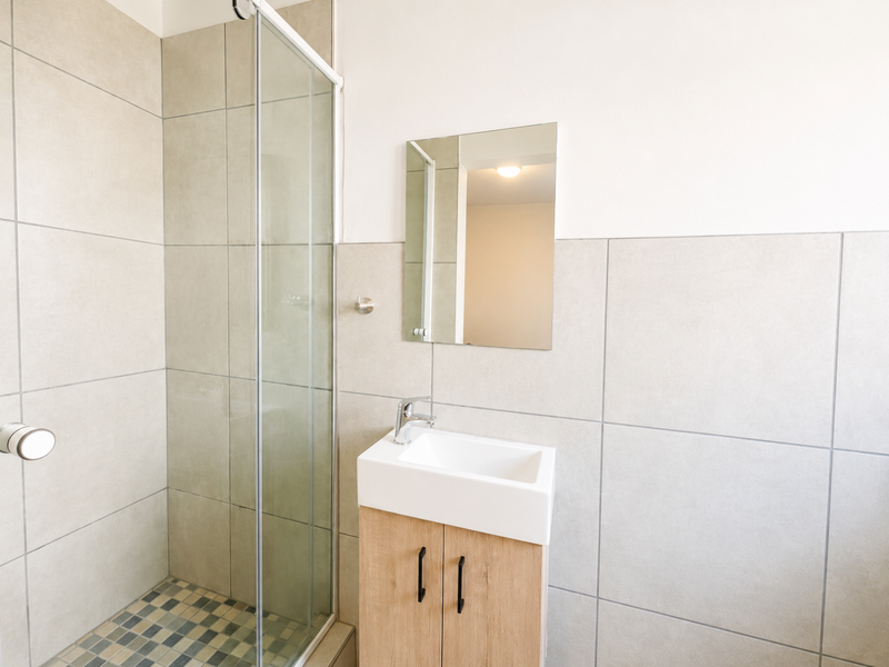 A modern, clean bathroom featuring a glass-enclosed shower, a compact vanity with a ceramic basin, and neutral wall tiling.