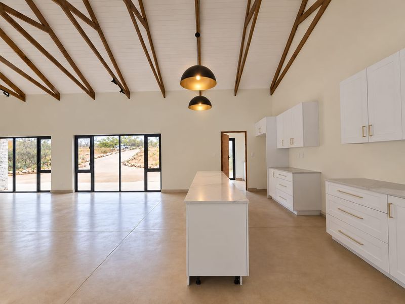 Spacious open-plan living and kitchen area featuring vaulted ceilings with exposed timber rafters, modern white cabinetry, stone countertops, and large windows with scenic views.
