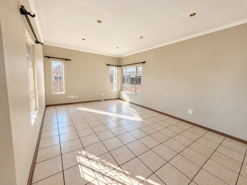 A bright, empty living room featuring neutral tiled flooring, recessed downlighting, and security bars on the windows.