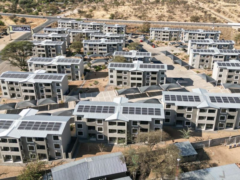 Aerial view of a modern apartment complex featuring multiple residential blocks, solar panel installations, paved parking areas with shade structures, and perimeter security fencing.