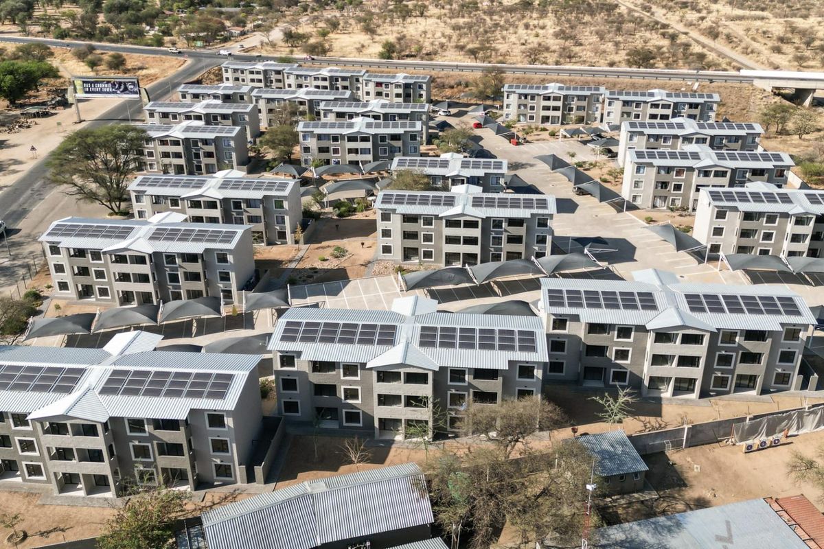 Aerial view of a modern apartment complex featuring multiple residential blocks, solar panel installations, paved parking areas with shade structures, and perimeter security fencing.