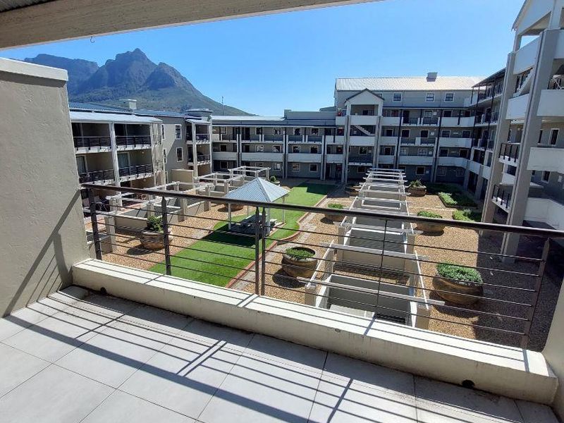 A spacious covered balcony with tiled flooring and metal balustrades, offering an elevated view of the apartment complex courtyard and surrounding mountain landscape.
