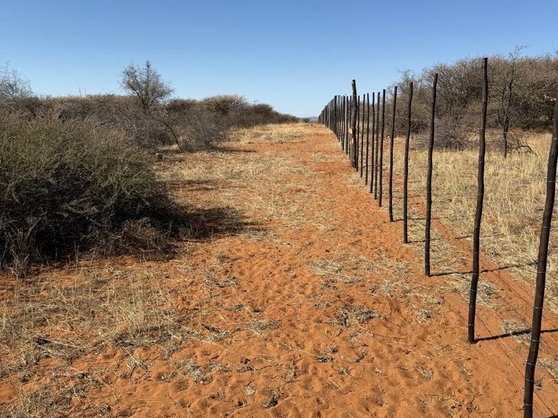 A view of an agricultural land plot featuring sandy terrain and a long stretch of wire fencing with wooden posts.