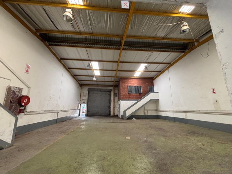 Spacious industrial warehouse unit featuring high ceilings with exposed steel trusses, concrete flooring, a roller shutter door, and an internal staircase.