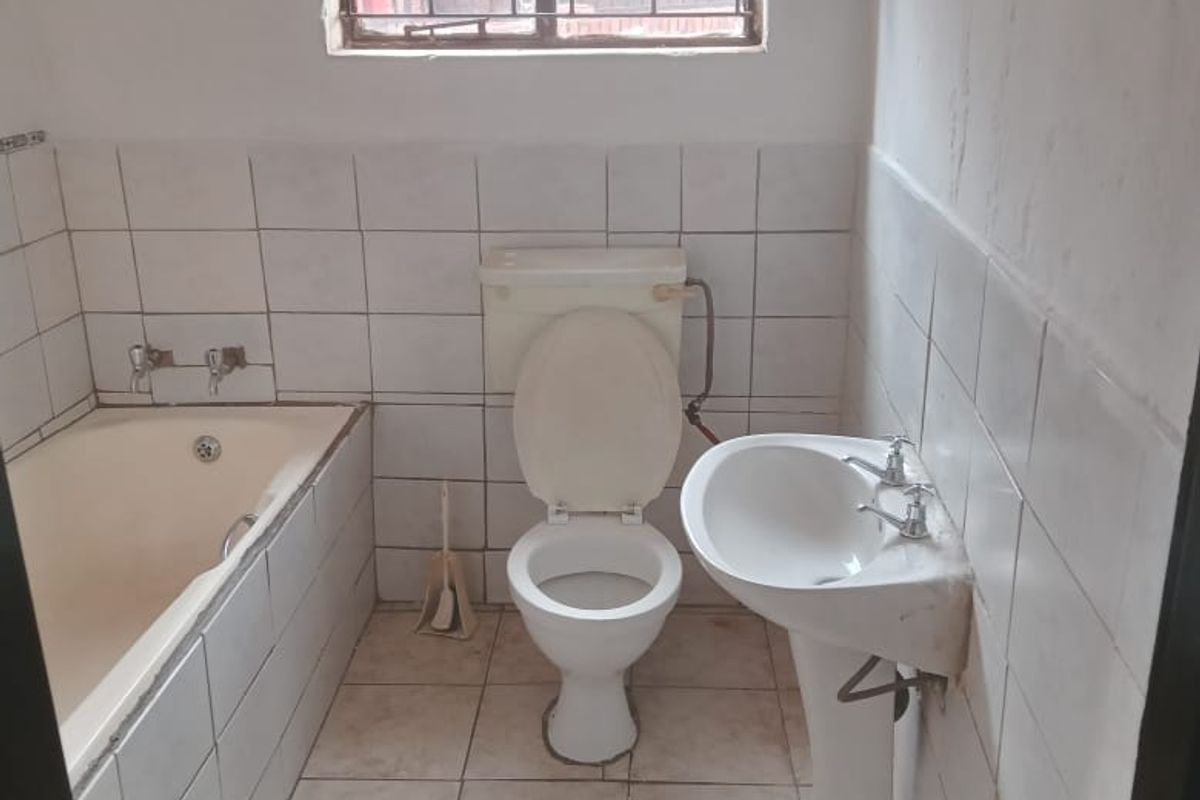 A dated bathroom featuring a standard bathtub, pedestal basin, and toilet, with tiled walls and flooring and a security-barred window.
