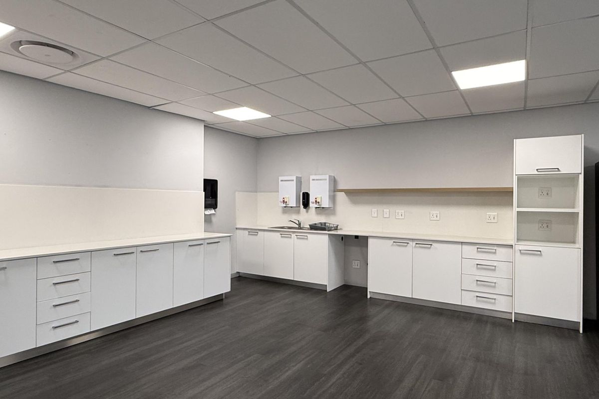 A clean, modern office kitchenette featuring white built-in cabinetry, laminate countertops, a stainless steel sink, and durable dark vinyl flooring.