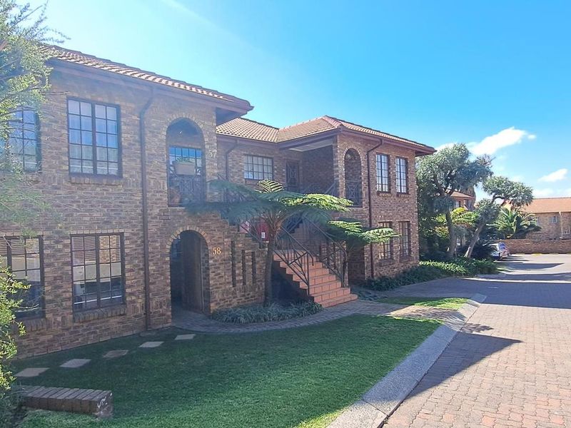 A well-maintained face brick apartment complex featuring arched entryways, an external staircase, and a paved driveway with landscaped garden areas.