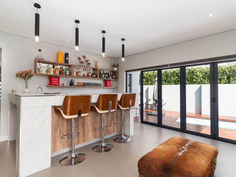 Modern indoor bar area featuring a stone-topped counter, integrated sink, floating shelves, and seamless access to the outdoor pool deck through bi-fold doors.