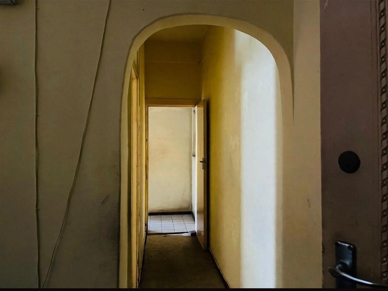 A narrow interior hallway featuring an arched doorway and dated wall finishes, leading to an adjacent room with tiled flooring.