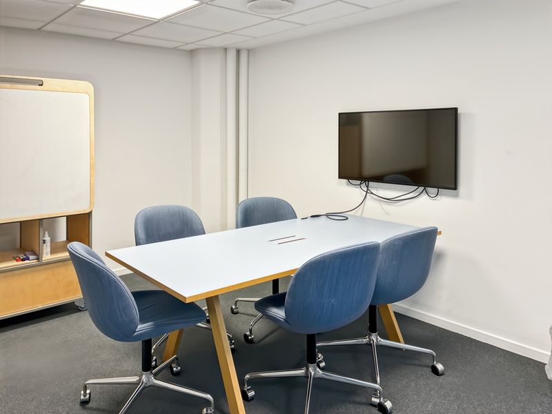 A functional office meeting room featuring a suspended ceiling, recessed lighting, wall-mounted display screen, and carpeted flooring.