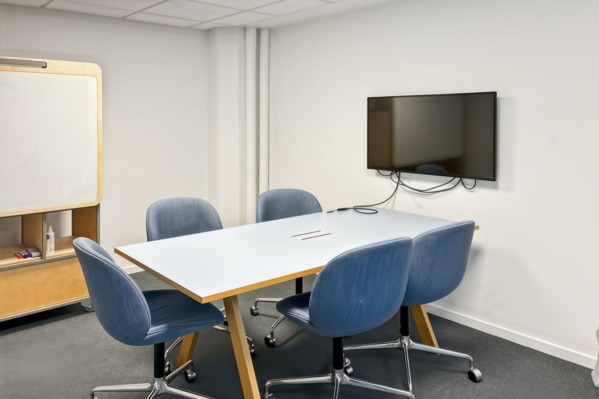 A functional office meeting room featuring a suspended ceiling, recessed lighting, wall-mounted display screen, and carpeted flooring.