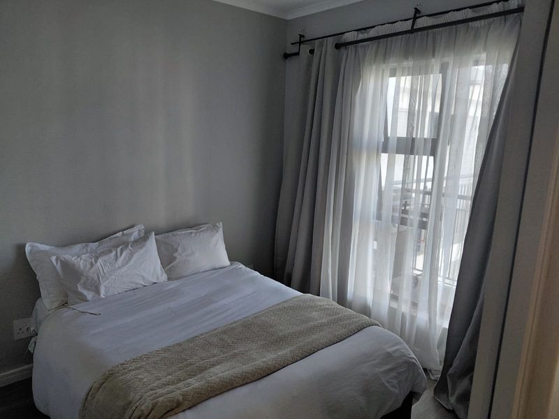 A cozy bedroom featuring neutral wall tones and a large window with layered curtains, providing natural light and a view of the exterior balcony.
