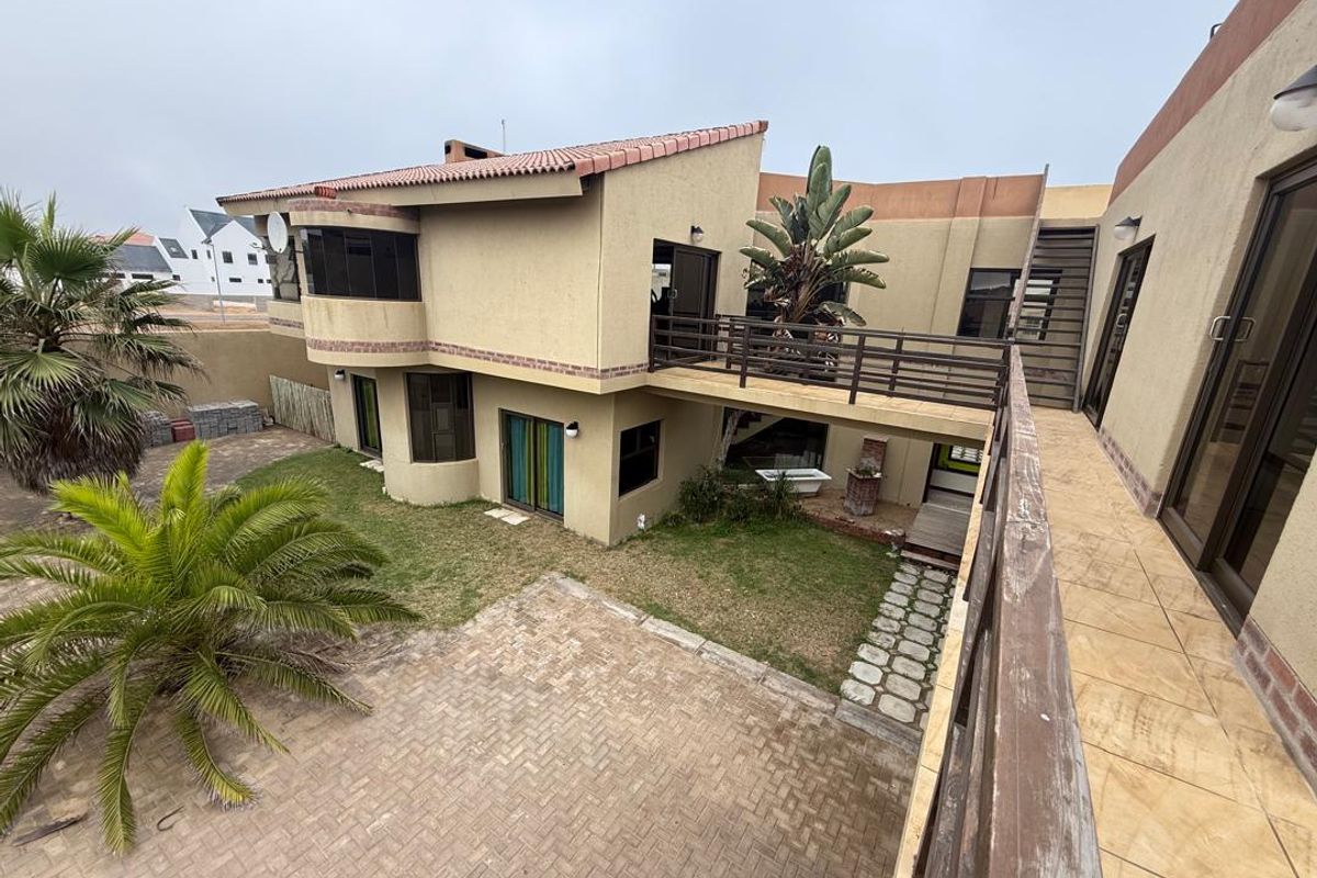 A rear exterior view of a multi-level house featuring a raised wooden walkway, paved courtyard, and landscaped garden area.