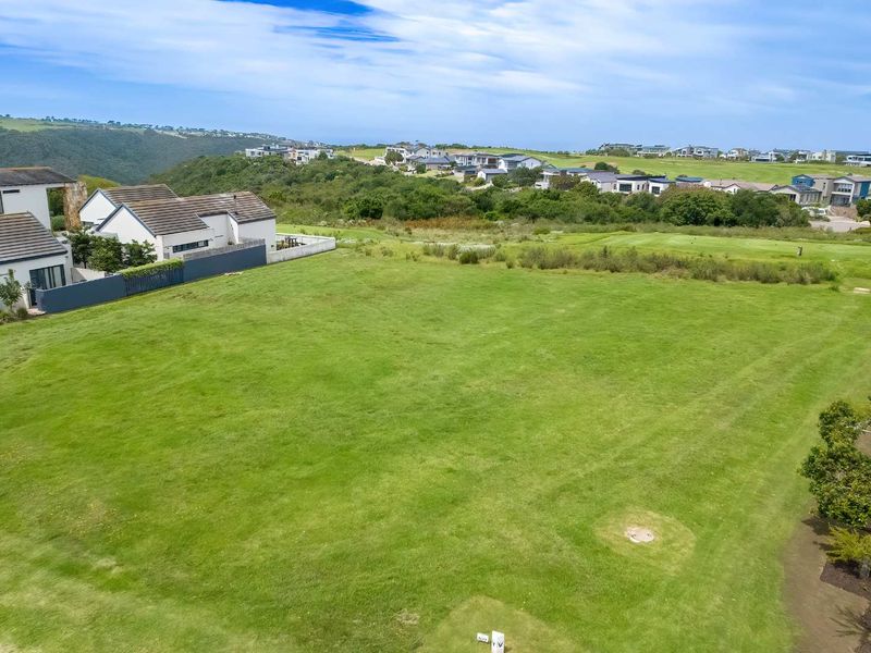 An elevated aerial view of a vacant residential plot featuring well-maintained grass, adjacent to existing residential properties and scenic landscape views.