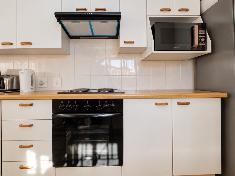 A well-maintained kitchen featuring white cabinetry, a wooden countertop, tiled backsplash, and integrated appliances including an oven, hob, extractor fan, and microwave.