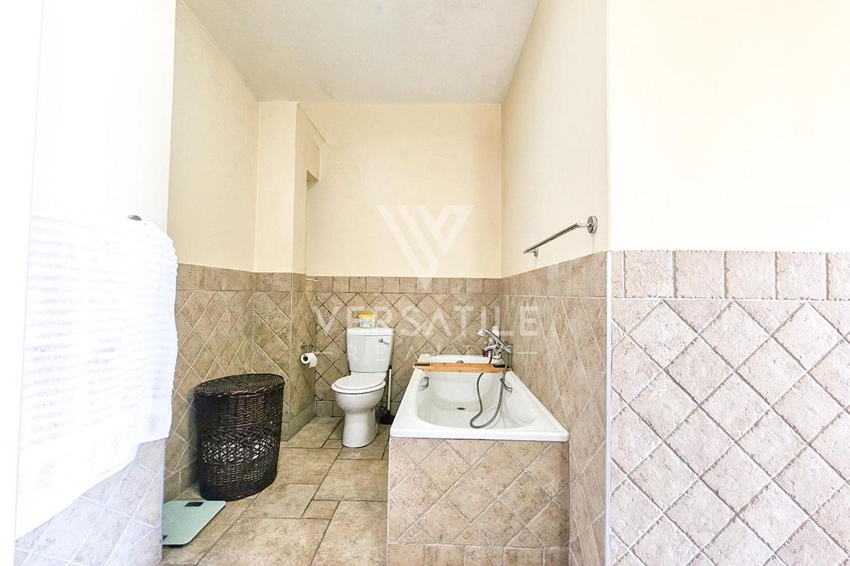 A functional bathroom featuring a built-in bathtub with shower mixer, a standard toilet, and neutral wall and floor tiling.