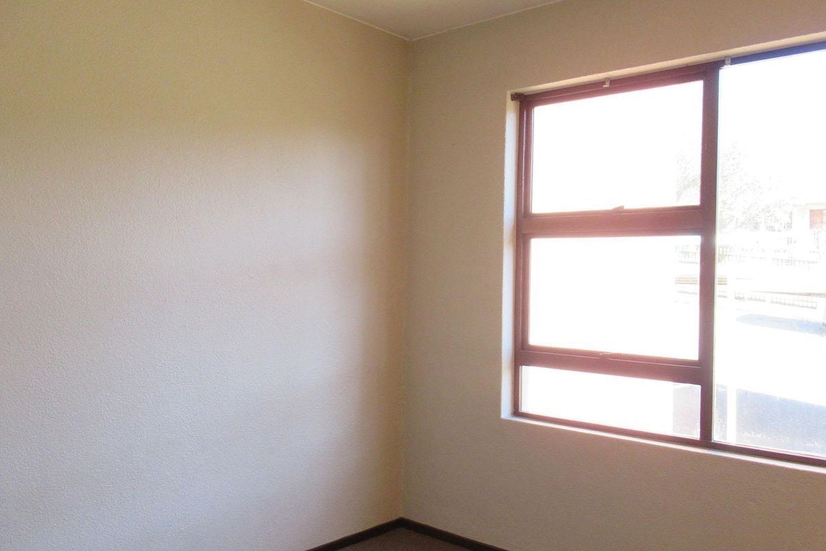A simple, empty bedroom featuring neutral walls and a large window with a brown frame, providing natural light.