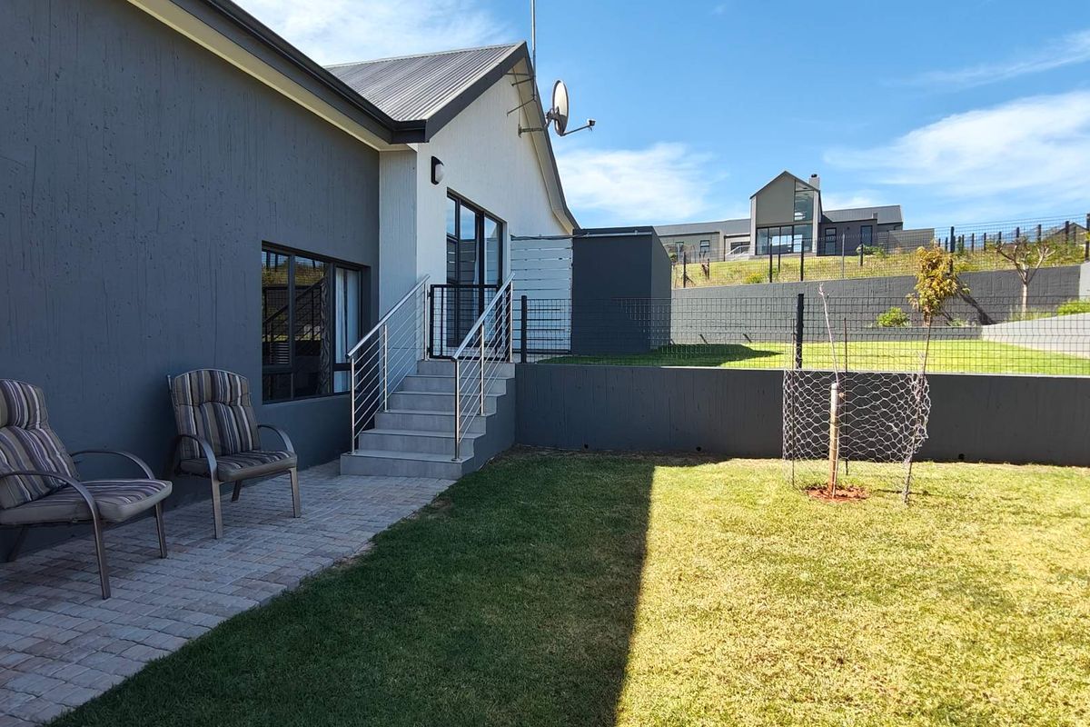 A well-maintained rear garden featuring a paved patio area, concrete stairs with stainless steel railings, and a landscaped lawn with perimeter fencing.