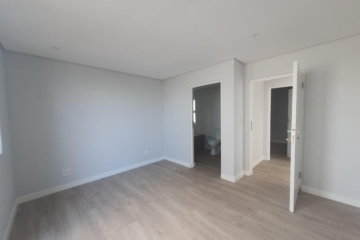 A bright, modern bedroom featuring neutral grey walls, light wood-look laminate flooring, and direct access to an en-suite bathroom.
