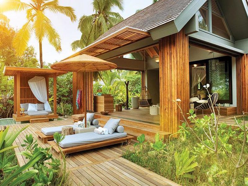 A luxurious tropical villa patio featuring extensive timber decking, natural wood cladding, and seamless indoor-outdoor flow through large sliding glass doors.