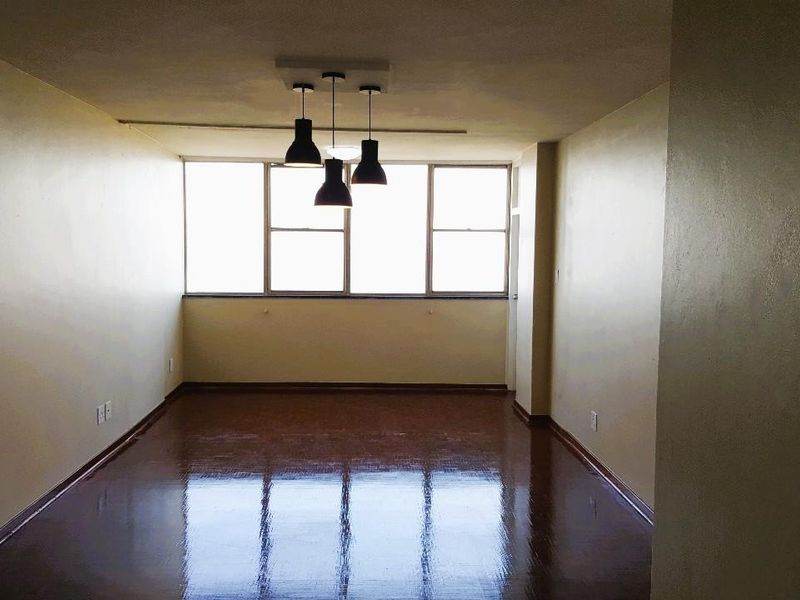 A spacious, empty living area featuring polished vinyl flooring, neutral walls, and a modern three-light pendant fixture, illuminated by a large window.