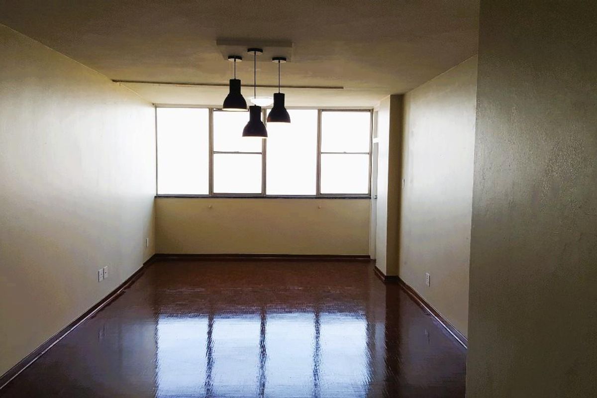 A spacious, empty living area featuring polished vinyl flooring, neutral walls, and a modern three-light pendant fixture, illuminated by a large window.