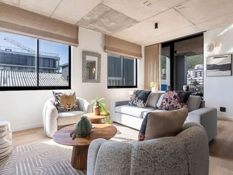 Modern living room featuring an exposed concrete ceiling, light hardwood flooring, and large windows providing ample natural light and access to a balcony.