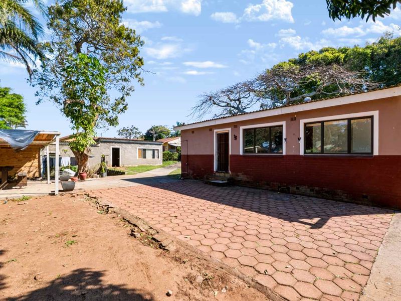 Rear exterior view of the property featuring a paved driveway, brick facade, and surrounding garden area with mature trees.