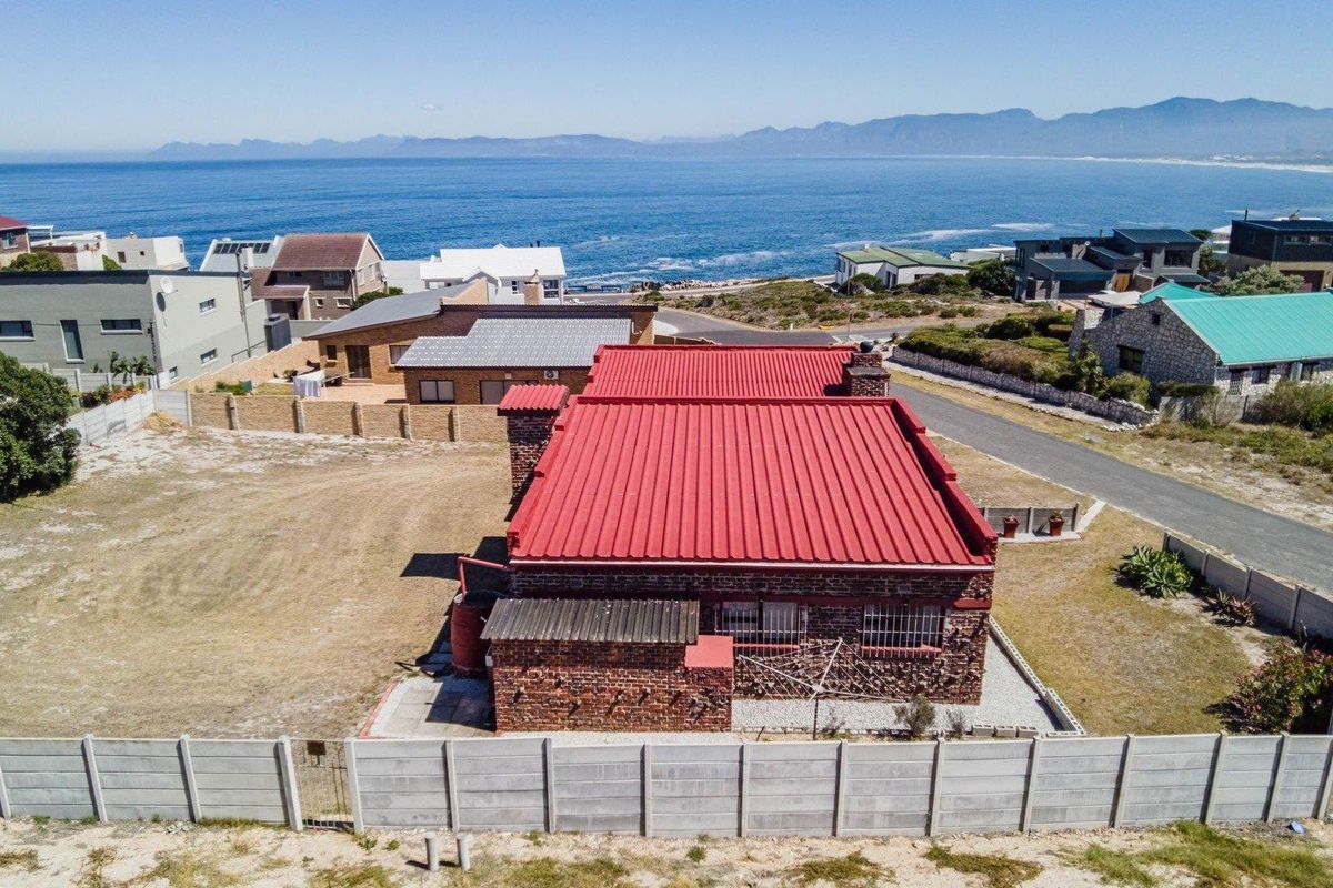 Aerial view of a coastal property featuring a red metal roof, brick exterior, and perimeter walling, set against a scenic ocean backdrop.