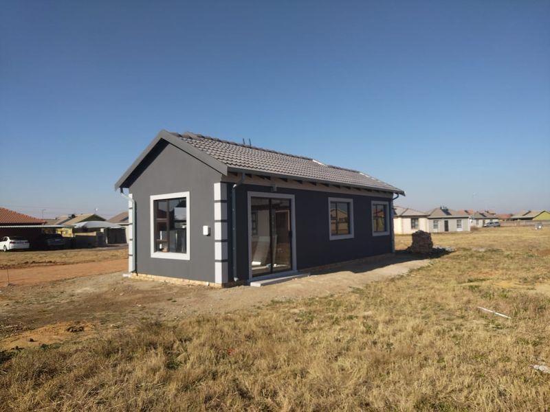A modern, newly constructed single-story house featuring a dark grey exterior finish, tiled roof, and aluminium-framed windows, situated on an undeveloped plot.