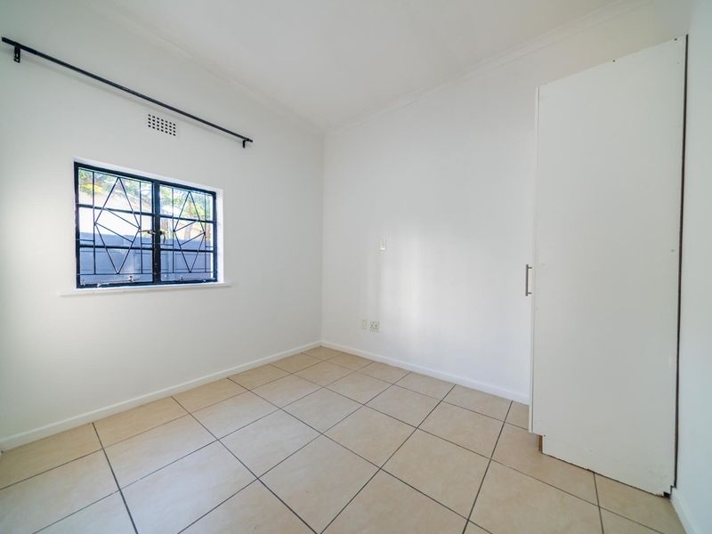 A clean, empty bedroom featuring neutral tiled flooring, a built-in cupboard, and a window fitted with security bars.