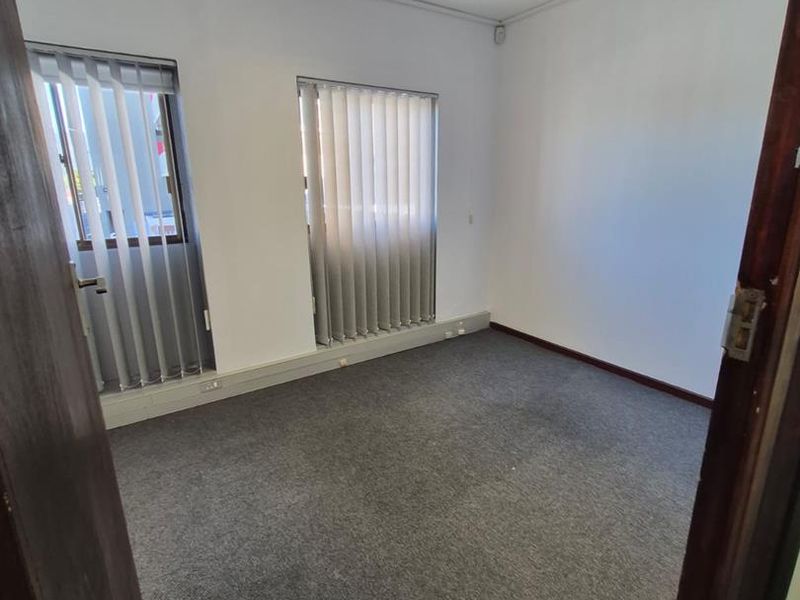A vacant office space featuring grey carpet flooring, vertical window blinds, and a ceiling-mounted fluorescent light fixture.