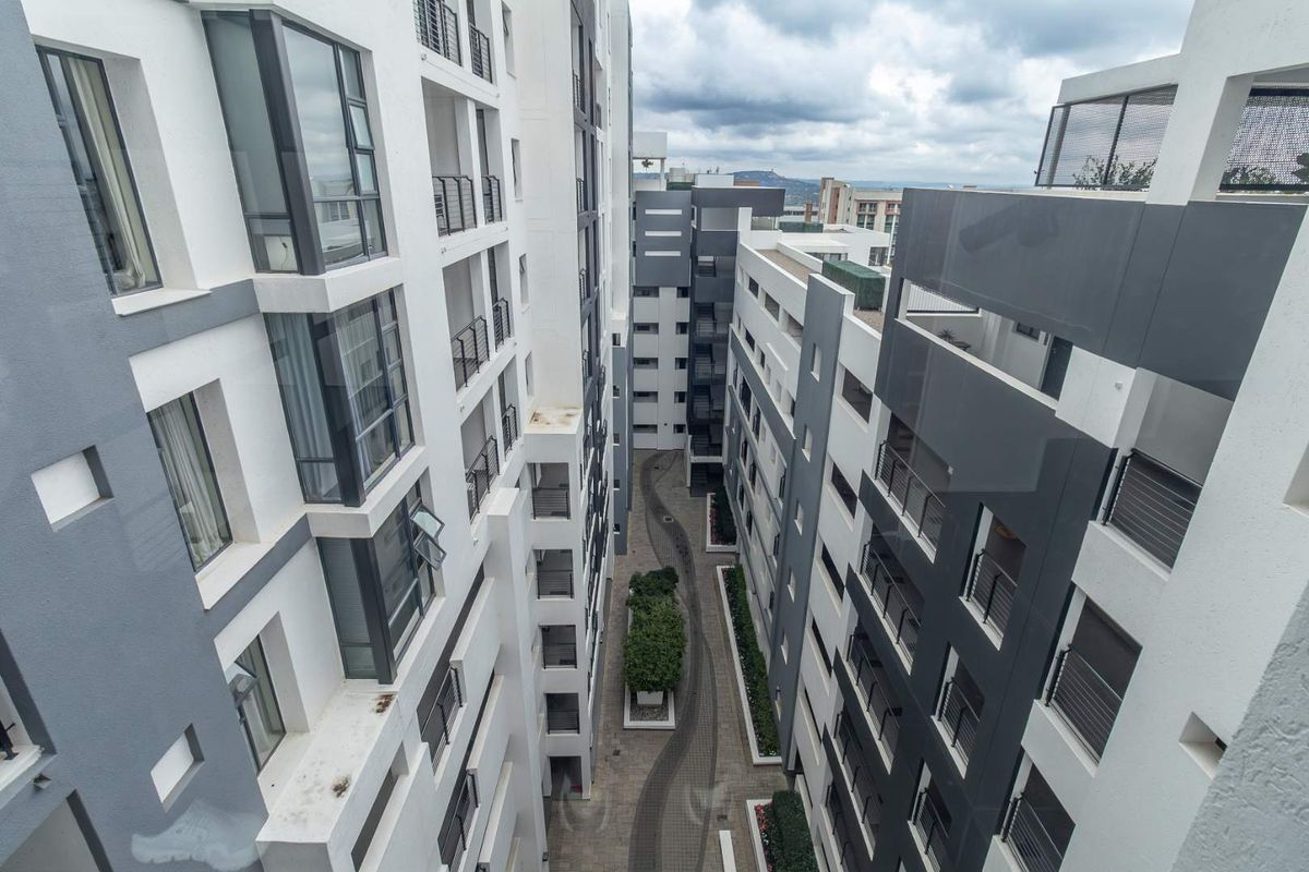 An elevated aerial view of the modern apartment complex, showcasing the building facades, balconies, and the central paved courtyard with landscaped garden features.