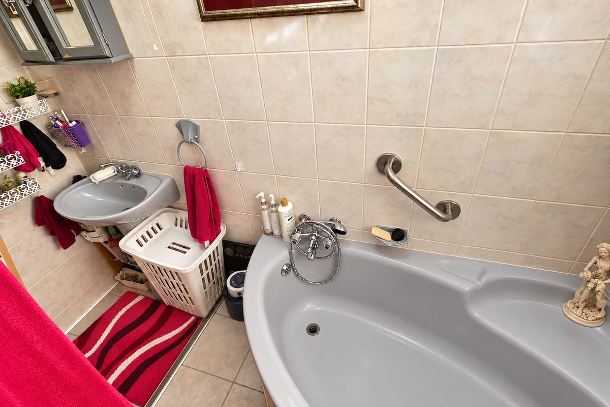 A functional bathroom featuring a corner bathtub, pedestal basin, and wall-mounted grab rail, finished with neutral ceramic wall and floor tiles.