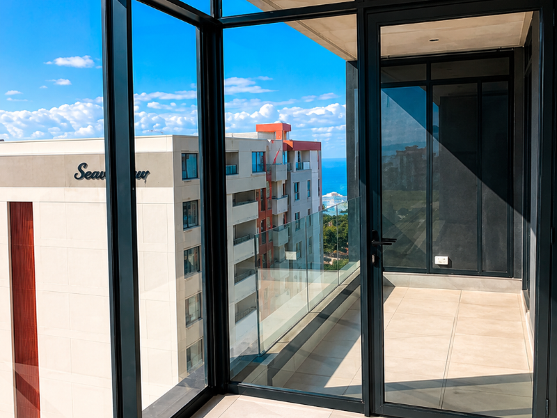 A modern, enclosed balcony featuring large glass panels, sleek aluminium frames, and neutral floor tiles, offering clear views of the surrounding urban landscape.