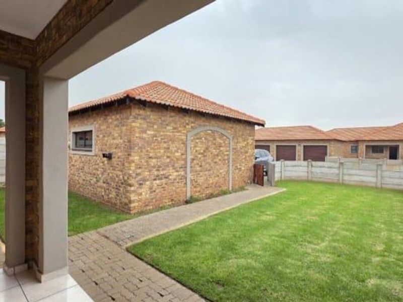 A view of the rear exterior featuring a covered patio, paved walkways, a well-maintained lawn, and brick-faced outbuildings enclosed by precast boundary walls.