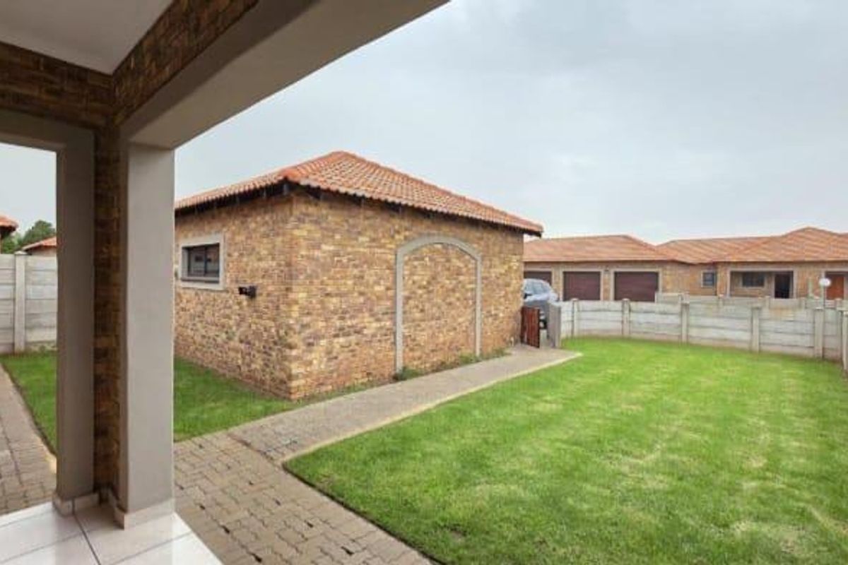 A view of the rear exterior featuring a covered patio, paved walkways, a well-maintained lawn, and brick-faced outbuildings enclosed by precast boundary walls.