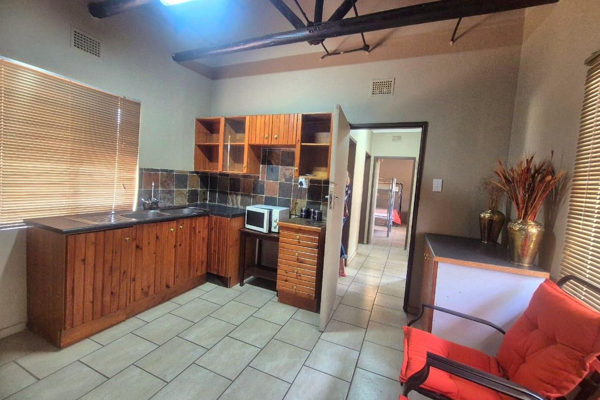 A functional kitchen area featuring wooden cabinetry, dark countertops, tiled flooring, and exposed timber roof trusses, with an adjacent hallway leading to bedrooms.