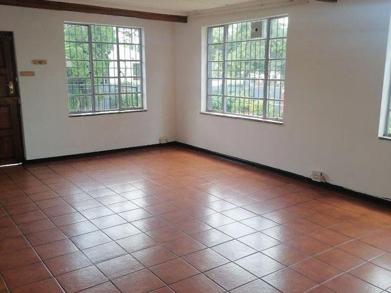 A spacious, empty living area featuring ceramic floor tiles, exposed timber ceiling beams, and large steel-framed windows with security bars.