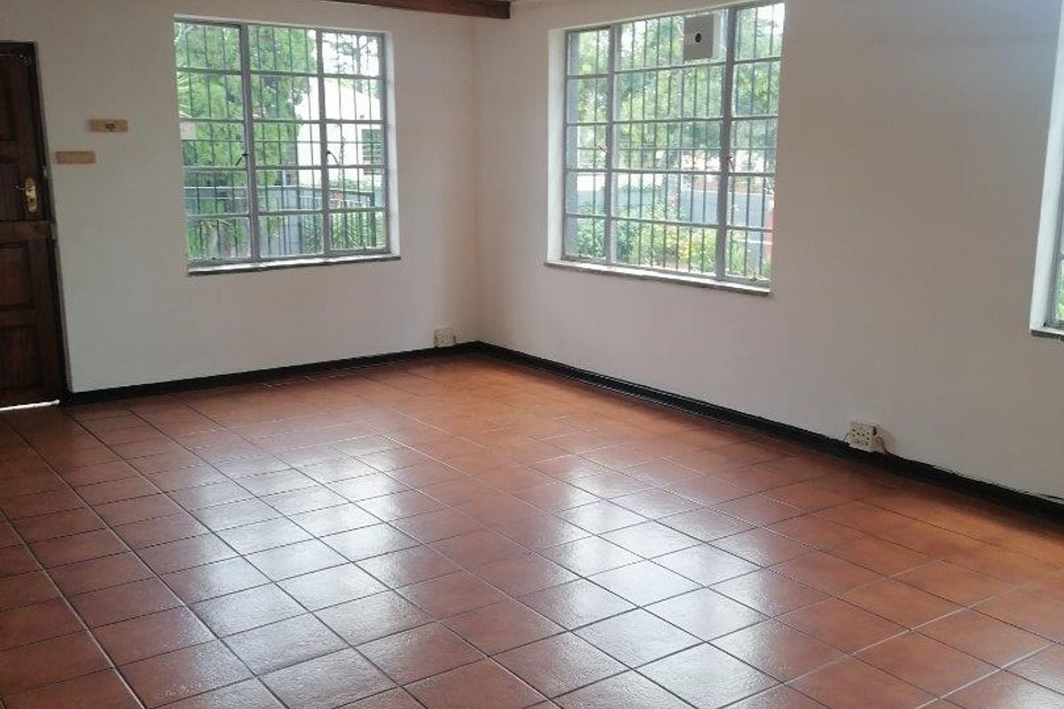 A spacious, empty living area featuring ceramic floor tiles, exposed timber ceiling beams, and large steel-framed windows with security bars.