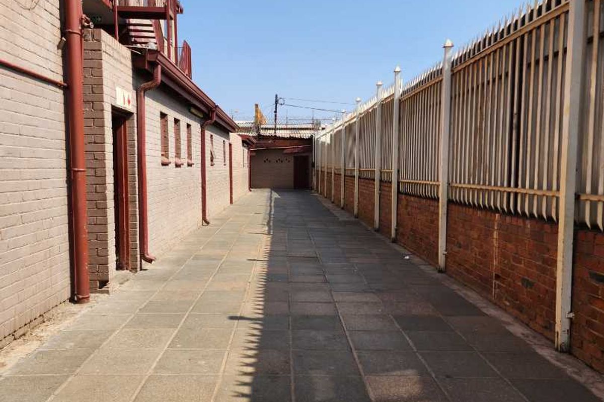 A narrow exterior walkway between the apartment building and a perimeter security fence, featuring paved flooring and metal security spikes.