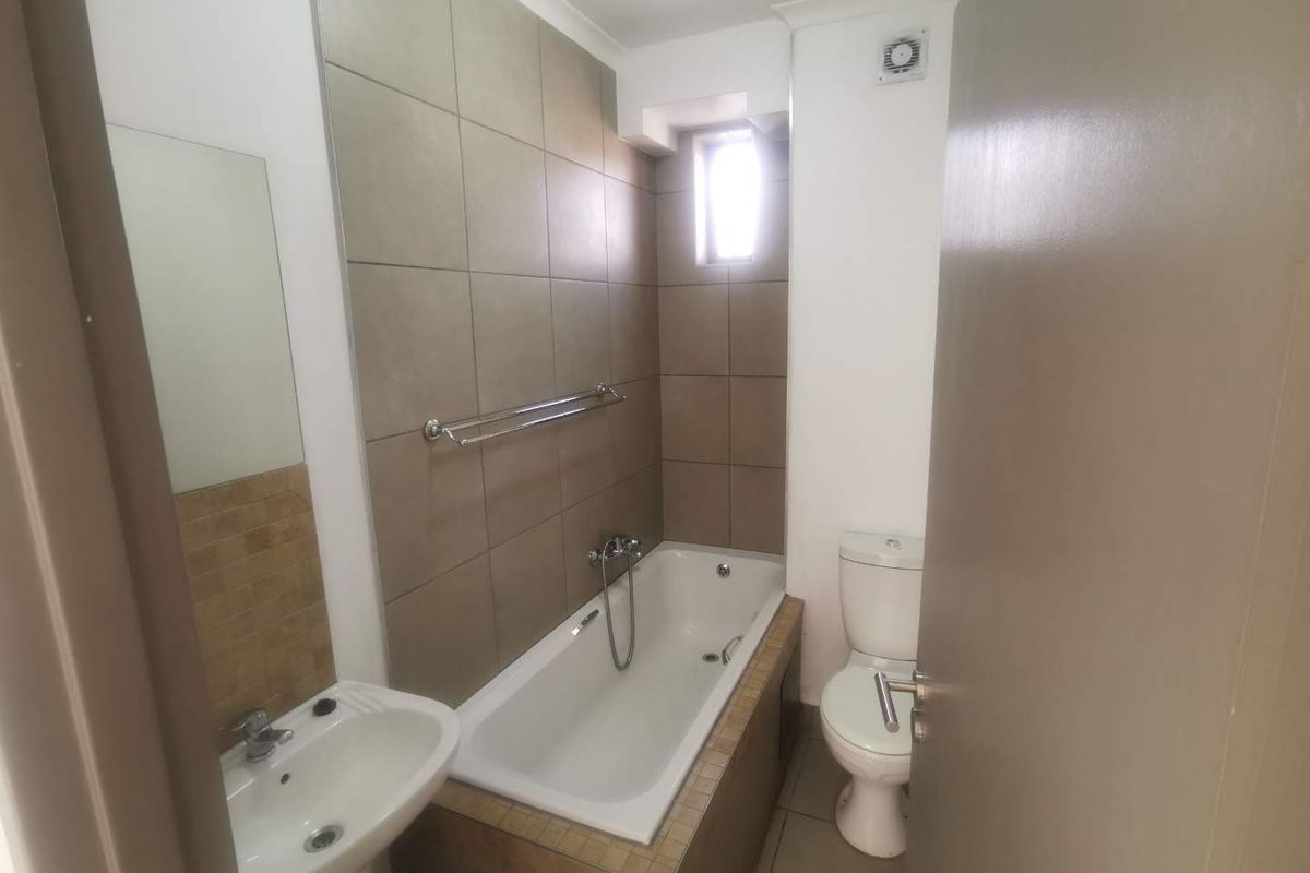A functional bathroom featuring a built-in bathtub with shower mixer, pedestal basin, and toilet, finished with neutral wall and floor tiling.