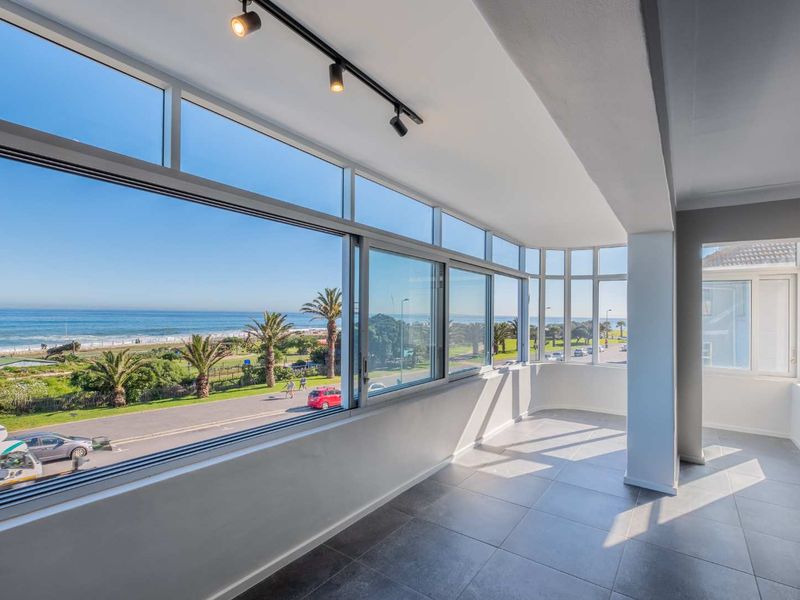 A bright, enclosed balcony featuring large sliding windows with expansive ocean and coastal views, finished with modern tiled flooring and track lighting.