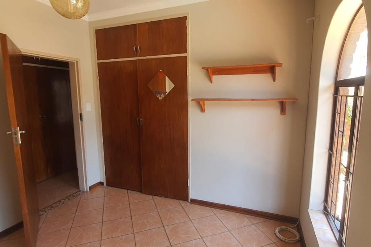 A bedroom featuring built-in wooden cupboards, tiled flooring, and an arched window with security bars.