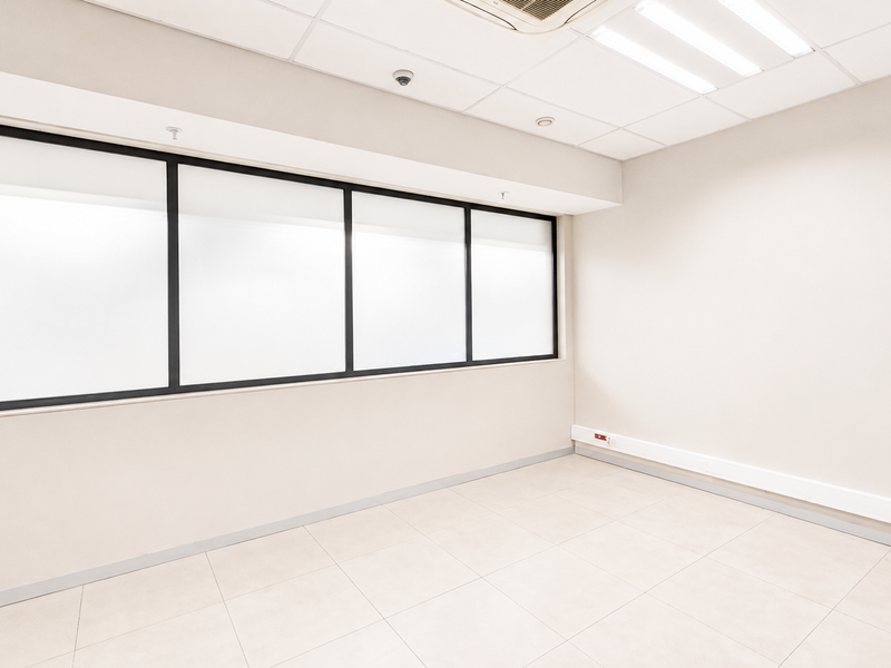 A bright, empty office space featuring tiled flooring, a suspended ceiling with integrated lighting, and a large window with a black frame.