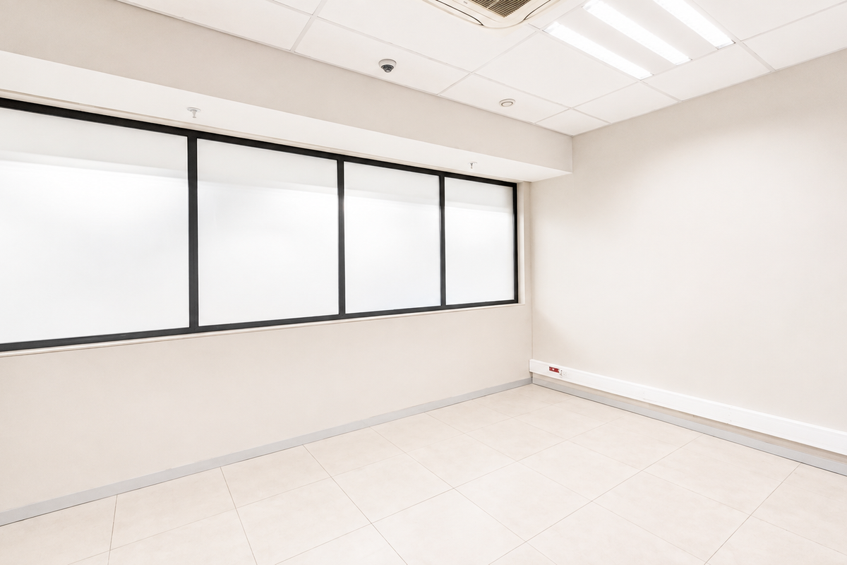 A bright, empty office space featuring tiled flooring, a suspended ceiling with integrated lighting, and a large window with a black frame.