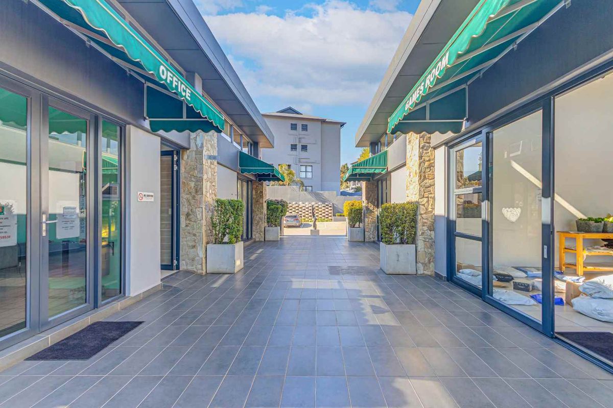 A well-maintained outdoor walkway between building wings featuring stone-clad pillars, tiled flooring, and green retractable awnings leading to a communal office and games room.
