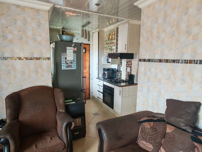 Open plan living area featuring a compact kitchen with built-in oven, hob, and extractor fan, finished with dated wall tiling and ceiling paneling.