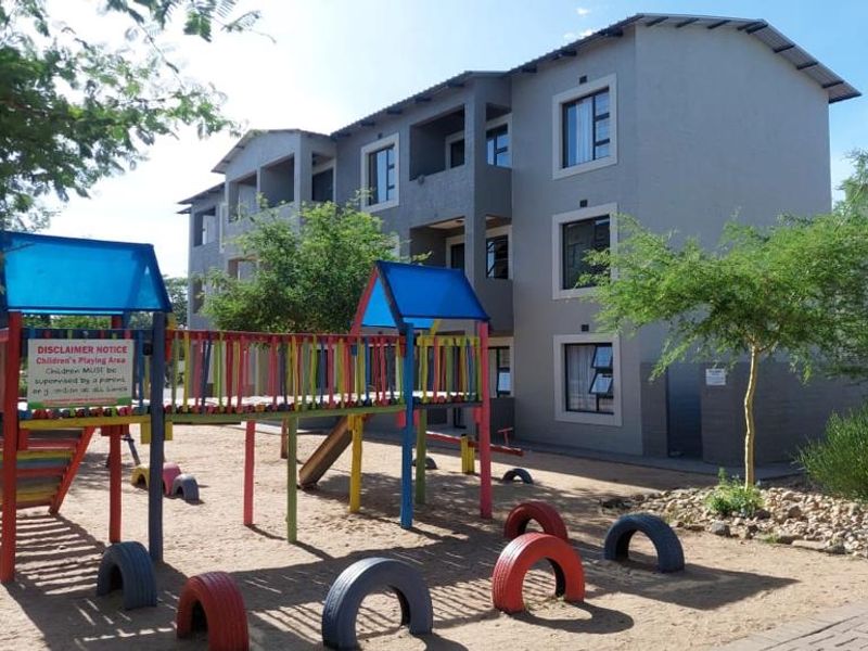 Exterior view of a multi-story apartment complex featuring a communal playground area with fixed play equipment and landscaped surroundings.