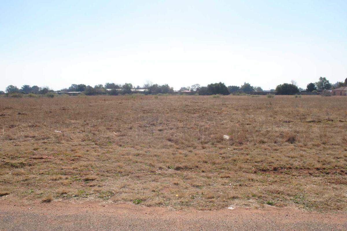 A wide-angle view of a flat, vacant residential plot of land with dry grass and surrounding vegetation.